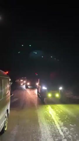 Here is a concise description of the video:

The video shows a convoy of vehicles driving on a wet, dark road at night. At least two fire trucks can be seen, including one with a flashing red light. Other vehicles, including a white van and a red pickup truck, are also present. The road is reflective, suggesting it may be wet. The overall scene indicates an emergency response or transportation convoy is traveling at night.