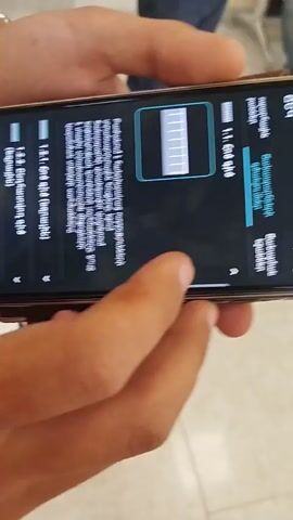 The video shows a close-up of a person's hands holding a smartphone. The phone's screen displays information in Armenian. The content on the screen appears to be a list of settings or menu options, indicated by numerical labels such as "1.1.1. CR8" and "1.3.2. CR9". There are also Armenian words and phrases visible, including "Կարգավորումներ" which translates to "Settings" or "Settings". In the upper right corner, a time stamp "10:32" is displayed. The phone screen also features a graphical element that resembles a battery indicator with multiple bars, and another graphic of a ruler or scale. The background of the video is blurred, but suggests an indoor setting with hints of people and possibly furniture. The hands holding the phone are those of an individual, with visible fingernails and skin texture. The lighting suggests it is daytime.