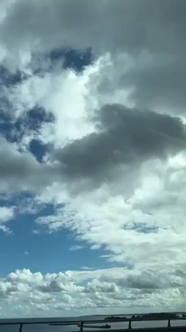 The video shows a view from a vehicle, likely a car, looking up at the sky. The sky is filled with a mix of white and dark gray clouds. There are patches of bright blue sky visible between the clouds. In the distance, at the bottom of the frame, a body of water is visible, possibly a lake or ocean. There appears to be a bridge or a series of structures crossing the water, and some distant land is also visible on the horizon. The camera is angled upwards, capturing the expansive sky. The lighting suggests it is daytime, possibly overcast with breaks of sunshine. There are no people or distinct objects in the foreground or mid-ground.