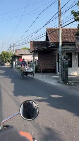 A person rides a bicycle with a cart attached. The cart is carrying plastic containers filled with a light-colored liquid. The person is wearing a blue shirt and dark pants. The cart is a two-wheeled vehicle with a metal frame and a wooden platform. The bicycle is a standard upright bicycle. The person is pedaling the bicycle and appears to be moving forward. The scene is a street in what appears to be a residential area. There are houses on either side of the street, and overhead power lines are visible. The sun is shining, and there are shadows on the street. The video is shot from the perspective of someone riding behind the bicycle.