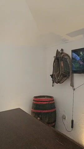 The video shows an interior scene. In the foreground, there is a dark wooden table and a bench. To the left, against a white wall, is a large wooden barrel with red bands. Mounted on the wall above the barrel is a horse's harness. To the right of the barrel, a television screen is mounted on the wall. Further to the right, a window is open, with bars on the exterior. Leaning against the wall next to the window is a large shovel and a pitchfork, both appearing to be made of wood and metal. On the wall to the left of the window, there is a wooden implement, possibly an agricultural tool, and a decorative panel with a painted grape vine design. Above the decorative panel is another large wooden agricultural implement, resembling a yoke or harness. The walls are white, and the ceiling is angled. The floor appears to be paved with stones. The video pans slightly to the right, showing more of the window and the items on the wall. Light streams in from the open window, illuminating the scene.