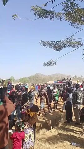 The video shows a crowd of people standing on a dusty, sunlit ground. Most individuals are adults, but several children are also present, some being carried. They are dressed in diverse, casual clothing. In the background, partially obscured by the crowd and dust, a black vehicle with flashing blue and red lights can be seen. People are moving around, pointing, and looking towards the vehicle. The atmosphere seems busy and somewhat excited. A tree branch extends into the frame from the top left, adding a natural element to the scene. The weather appears to be sunny with a clear, blue sky.