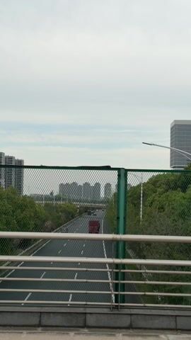 The camera is static, positioned on a bridge, overlooking a multi-lane highway. A chain-link fence with green metal posts and a silver metal railing are in the foreground. Cars and trucks are visible on the highway, moving away from the camera. Green trees line the sides of the highway. In the background, tall buildings are visible against a cloudy sky. The weather appears overcast.