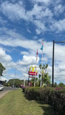 The video shows a McDonald's sign with the iconic golden arches, along with two flags flying on poles. One flag is red and the other is blue with a Union Jack in the corner. The McDonald's sign has the words "McDonald's Drive-Thru" visible. There are palm trees and other greenery in the background, and the sky is blue with white clouds. The overall scene suggests a sunny day at a McDonald's restaurant.