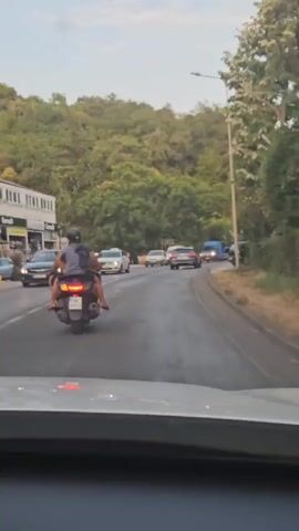 Two people, a driver and a passenger, are riding on a scooter on a paved road. Both are wearing helmets. The driver is wearing a grey t-shirt and black shorts. The passenger is wearing a grey t-shirt and black shorts. They are both barefoot. The scooter has a black license plate with white lettering that reads HN AH49. The scooter has its brake lights on. The road is a two-lane road with traffic in both directions. There are cars and a van on the road. On the left side of the road, there is a building with several businesses and some outdoor market stalls. Behind the businesses, there is a hill covered in trees. The sky is overcast. The video is shaky, suggesting it was filmed from a moving vehicle.