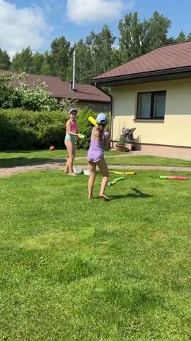 Two young girls are playing in a grassy yard on a sunny day. The girl on the left is wearing a pink swimsuit and hat and holding a green water gun. The girl on the right is wearing a purple swimsuit and blue hat and is also holding a green water gun. There are several other water guns scattered on the grass. In the background, there are houses and trees. The girl on the right appears to be filling her water gun from a white bucket.