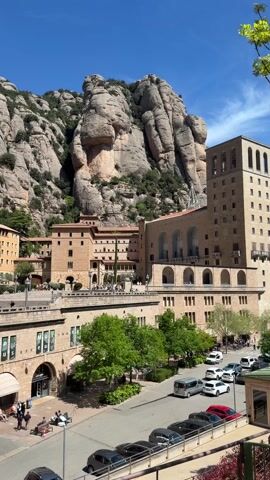 The video shows the Montserrat Monastery complex in Spain, set against the backdrop of the Montserrat mountain range. The buildings of the monastery are constructed from light brown stone, and include arched walkways, a tall tower, and a museum with "Museu" signs displayed. Cars are parked along a road in front of the complex, and people are seen walking and sitting on benches in the plaza. The mountain range behind the monastery features distinctive, rounded rock formations and sparse vegetation. The sky is clear and blue.
