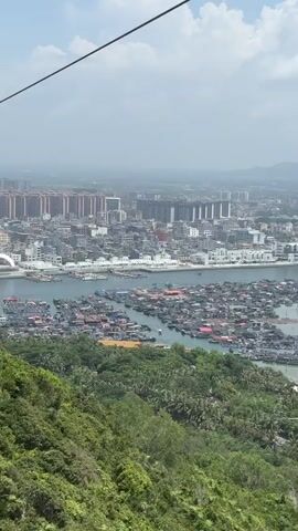 An aerial drone shot pans across a coastal cityscape and bay. In the foreground, lush green trees cover a steep hillside, with a thin cable visible at the top left of the frame. The camera moves right and slightly upward, revealing a bay filled with numerous small boats and floating structures packed closely together, resembling a village on the water. Beyond the bay, a city stretches out, characterized by a dense cluster of buildings, including taller, modern structures and smaller, more uniform residential blocks. A waterfront promenade or road runs along the city's edge. Further in the distance, the landscape transitions to rolling hills and a hazy horizon, indicating a sprawling urban area. The sky is partly cloudy with bright sunlight illuminating the scene.