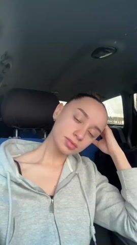 A young woman with her hair pulled back in a bun, wearing a grey hooded sweatshirt, is seated inside a car, likely in the passenger seat. The car interior features blue and black seat covers. She looks directly at the camera throughout the video. Initially, her right hand is near her chin with fingers slightly curled. She then changes her pose, resting her chin on her right hand, showcasing white nail polish. The background outside the car appears to be a bright daytime setting with indistinct views of buildings or trees. Her expression is neutral and calm. There is no visible text. The video was filmed in Khanskaya, Russia.