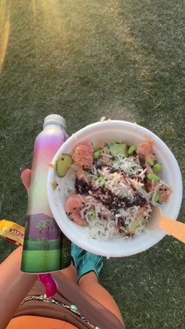 The video shows someone holding a bowl of food, possibly a poke bowl, and a water bottle at an outdoor event. The bowl contains rice, salmon, edamame, cucumber, and what appears to be some sort of sauce or topping. The water bottle is a colorful, iridescent design. The camera then pans out to reveal a grassy field filled with people. Palm trees are visible in the background, and the sun is setting. There is a stage or structure in the distance, suggesting a concert or festival. The crowd is casually dressed, many in festival attire. Overall, the scene captures a vibrant outdoor event during sunset.