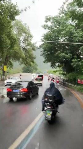 The video shows a rainy day on a road with cars and motorcycles. The camera appears to be mounted on a motorcycle moving along the road. The road has white lines separating the lanes. There are other motorcycles and cars ahead, some with their brake lights on.  The person on the motorcycle is wearing a rain poncho. Trees line the side of the road. In the distance, there is a mountain obscured by fog. A security camera is visible above the road. The sky is overcast and gray.  The road is wet from the rain, causing reflections of the vehicles' lights.