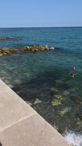 The video shows a bright, sunny day with a clear blue sky. The camera pans across a coastal scene. In the foreground, a paved walkway runs alongside the ocean. The water is a clear turquoise, with waves gently breaking on a rocky shore. A concrete barrier separates the walkway from the water. In the distance, a cliff face rises steeply, dotted with buildings and trees. Cars and a few pedestrians are visible along a road that runs parallel to the walkway. The overall impression is one of a scenic seaside location, likely in a Mediterranean climate.