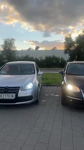 A silver Volkswagen Golf is parked in a paved lot. The car's headlights are on. In the background, there are trees, buildings, and a cloudy sky. Another car is parked further away. The camera then pans to the left, showing a street with grass and trees. A blue car is parked, and a person is bending down near its open trunk. The camera pans further left, showing a utility pole and more trees and buildings. The video ends.
