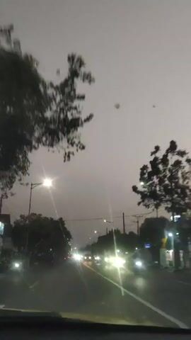 The video shows a view from a car driving on a road at dusk or dawn. The sky is overcast and gray. There are streetlights on both sides of the road, and the headlights of oncoming vehicles are visible. Trees line the sides of the road, and there are some utility poles and wires overhead. The road appears to be wet, suggesting it may have rained recently. Traffic is light to moderate, with cars and a truck visible. The camera is focused on the road ahead, and the scene is somewhat blurry.