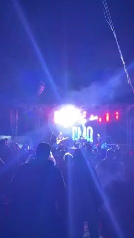 An outdoor night shot captures a rock concert with a band performing on a brightly lit stage, viewed from behind a silhouetted audience. The stage features four musicians, including two guitarists and a drummer, illuminated by red, blue, and white stage lights. A large LED screen behind the band displays changing graphics, including text like "EMO" and a stylized skull logo. Metal trusses frame the stage structure. The foreground shows the backs of numerous audience members, mostly in silhouette, with some raising their hands. A striped banner hangs in the upper left background against the dark night sky.