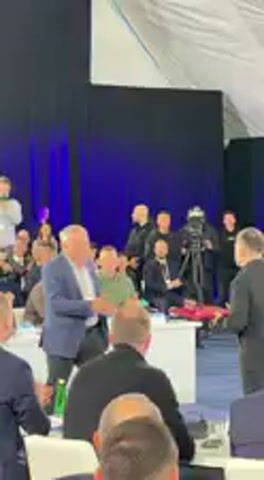 In an indoor setting, likely a conference hall or event space, two men are engaged in a formal exchange. The man on the left, an older individual wearing a light blue blazer over a white shirt and dark pants, is handing a red folder or plaque to the man on the right. The man on the right, younger and dressed in a black collared shirt and dark pants, accepts the item and shakes hands with the older man. Both men appear to be smiling.

Behind them, a group of people stands, some of whom are holding cameras, indicating a media presence. In the foreground, other attendees are seated at white-clothed tables, observing the event and applauding. The backdrop consists of a dark blue curtain or wall, with a white tent-like structure visible above. The lighting is artificial, illuminating the scene. The overall atmosphere suggests an award ceremony, official presentation, or similar public event taking place in Barvinok, Ukraine. No specific text is visible on the exchanged item or in the background.