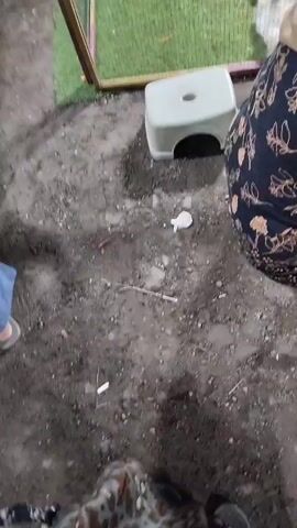 A ground-level view of several guinea pigs in an enclosure. The enclosure has a wooden frame and green artificial turf inside. There are at least five guinea pigs visible, varying in color from white to brown and black. One person's legs are visible on the left side of the frame wearing blue jeans and a sandal. Another person is partially visible on the right, wearing a dark patterned garment. A light gray plastic stool is positioned in front of the enclosure. The ground outside the enclosure appears to be dirt or gravel.