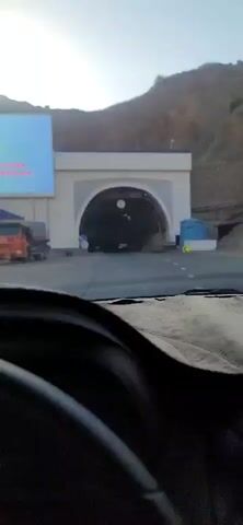The video shows a view from inside a car as it approaches and enters a tunnel. A large billboard with blue and white lettering is visible on the left side of the road. An orange truck is parked on the left. The tunnel entrance is arched and decorated with white patterns. Traffic signs indicating a speed limit of 50 and a directional arrow are visible near the tunnel. Inside the tunnel, construction materials and scaffolding are present. A height restriction sign indicating 4.5 meters is visible. The car's dashboard is visible in the foreground.