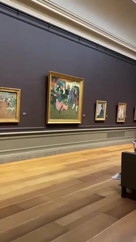 The video opens on a wide shot of a dimly lit art gallery with polished wooden floors. A large, dark brown wall dominates the upper portion of the frame, adorned with several framed paintings. The closest painting to the left is partially visible, depicting a vibrant scene. The centerpiece painting, centrally located, is a colorful depiction of people dancing. To its right, several other smaller paintings hang in a row, featuring portraits and more abstract scenes. In the foreground, a long, dark grey sectional sofa is positioned facing the paintings. Two individuals, one in a brown top and the other in a patterned dress, are seated on the sofa, engrossed in their mobile phones. As the camera pans to the right, more gallery visitors come into view. A person in a white t-shirt and shorts stands near a painting, holding a phone. Further to the right, a person wearing a hat and a striped shirt walks by, accompanied by a young girl in a blue dress and white sneakers. The gallery ceiling features a grid of skylights, illuminating the space with natural light. The overall atmosphere is quiet and contemplative, typical of a museum setting.