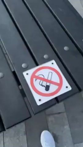 The video shows a bench with a "No Smoking" sign on it, then pans up to reveal a view of the Kelpies, two large horse head sculptures, in a park. The sky is cloudy and it appears to be dusk or early evening. There are picnic tables nearby. A few people can be seen in the distance.