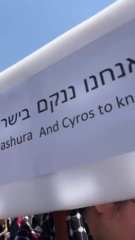 The video shows a close-up of a sign held by a person. The sign has text in both Hebrew and English. The English text reads "ashura And Cyros to know us." The person holding the sign is wearing dark clothing and a keffiyeh scarf around their neck. They appear to be in a crowd, with other people visible in the background and flags, some of which are red, green, and white. The scene seems to be outdoors under a clear blue sky. The person is holding a phone to their ear.