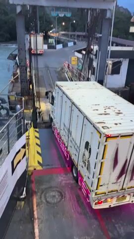 A white trailer truck with pink accents on the side and rear is slowly driving onto a ferry. The truck is positioned on a ramp that connects the ferry deck to the shore. The ferry deck is metal with yellow and black striped markings and red painted lines. The water is visible on the left side of the frame, and a mountainous landscape is in the background. Other vehicles and structures are visible on the shore. The sky is overcast. The video is filmed from a slightly elevated perspective, looking down at the truck.