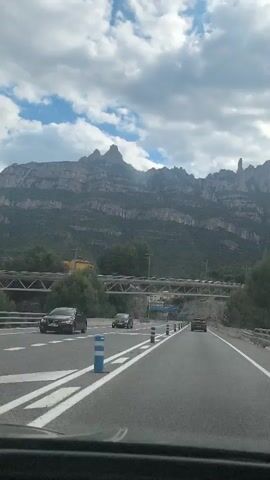 A first-person view from inside a vehicle moving on a multi-lane highway captures a striking mountain range in the background. The sky is partly cloudy, with patches of blue visible amidst the white clouds. The mountain range is rugged, with varying elevations and green vegetation covering its slopes. A bridge, constructed of metal beams and concrete supports, spans over the highway in the middle ground. The highway itself is marked with white lines, dividing the lanes and separating the direction of travel. There are blue and white bollards along the side of the road. Two cars are visible on the road, moving in the same direction as the camera. A construction sign is visible on the side of the road.