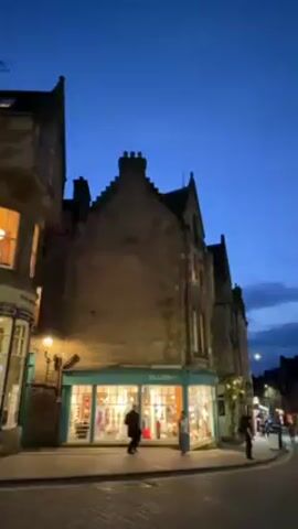 The video shows a cobblestone street in Edinburgh, Scotland, at twilight. The buildings are tall and made of stone, with steep gabled roofs and multiple windows, some of which are illuminated. A shop called "SWASH" is visible on the corner of a building, with bright light shining from its windows. Several people are walking on the street, and there are cars parked along the side. The sky is a deep blue, with some clouds visible. Overall, the scene depicts a quiet evening in a historic European city.