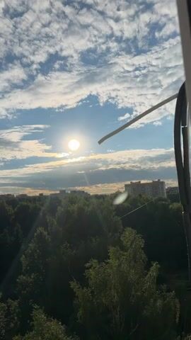 This is a vertical video showing a view from a window, looking out at a city skyline and a bright sky. The sun is high in the sky, creating lens flares and a general bright, warm atmosphere. The sky is blue with scattered white clouds, some of which are elongated and wispy.

Below the sky, a dense line of green trees stretches across the middle ground of the frame, their foliage appearing lush and healthy. Behind the trees, a row of tall, grey apartment buildings dominates the background. These buildings are typical of Soviet-era architecture, with many windows and balconies.

The sun's rays are casting a warm glow over the scene, suggesting it is late afternoon or early evening on a clear day. The presence of the trees and apartment buildings indicates an urban or suburban setting. The video is static, with no discernible activity taking place. The overall mood is peaceful and serene, bathed in the warm light of the setting sun. There is no visible text in the video.