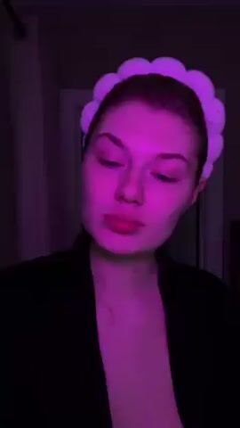 A woman with fair skin is illuminated by purple light as she sprays a product from a silver bottle onto her face. She is wearing a black robe or shirt and a white, fluffy headband. Her eyes are partly closed as she sprays the product. Music plays in the background. A white curtain and doorway are visible behind her.