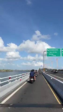 The video begins on a highway overpass, looking forward and slightly to the right. The sky is blue with scattered white clouds. To the left, water is visible with a bridge in the distance. To the right, traffic moves along the highway, and a green sign indicates "BANDARA NGURAH RAI KUTA 1 km". The road is paved black with yellow and white lane markings. A grey metal pole with a white railing separates the road from the edge of the overpass. Several motorcycles are on the road ahead, with people riding them. The camera appears to be mounted on a motorcycle or is following one, as the perspective shifts slightly, showing the back of a motorcycle with two riders in the foreground. The rider in the back is holding up a smartphone, possibly taking a picture or recording. The motorcycles continue forward on the highway.