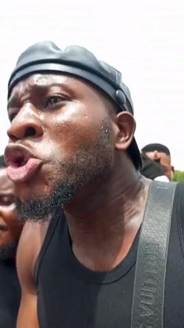 Here is a detailed description of the video:

The video features a man in a black tank top with the word “INFLUENCER” on the front, wearing a black hat and Louis Vuitton strap across his shoulder. He is surrounded by several people, some of whom are holding phones and filming him. A microphone is also visible. The man appears to be agitated and is shouting something. His skin is shiny with perspiration. The background includes a road and some greenery. The scene appears to be outdoors during the day.