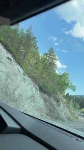 The video shows a view from inside a car driving on a paved road through a wooded area. The sun is shining and there are white clouds in a blue sky. Trees line both sides of the road, with a rocky embankment on the left. Several other cars are visible on the road ahead, and a guardrail runs along the right side of the road. A side mirror reflects the driver holding a phone. The overall impression is of a scenic drive on a sunny day.
