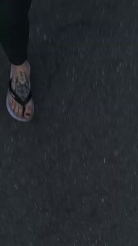 The video shows a top-down view of someone walking on an asphalt surface. The person is wearing dark green pants and flip-flops. One foot has a tattoo on the top of it. The camera is moving, creating motion blur.