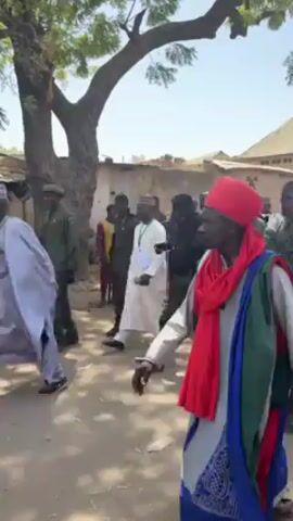 The video shows a procession down a dirt road in what appears to be a village or small town. The road is flanked by buildings and a large tree with bare branches. The procession is led by an older man wearing a red headwrap and a colorful robe, followed by a group of individuals in green military-style uniforms, some carrying weapons. Other people, dressed in civilian clothing, mingle among and follow the uniformed group. The camera follows the procession at a medium distance, maintaining the perspective of someone within or alongside the group. The overall atmosphere suggests an event of local significance or celebration.