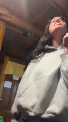 A young woman with long brown hair, wearing a gray hoodie and matching sweatpants, is indoors under a wooden ceiling, singing and dancing while looking up. She has a nose piercing and shows her teeth while smiling. The room has a warm, wooden interior, and a clock is visible in the background.