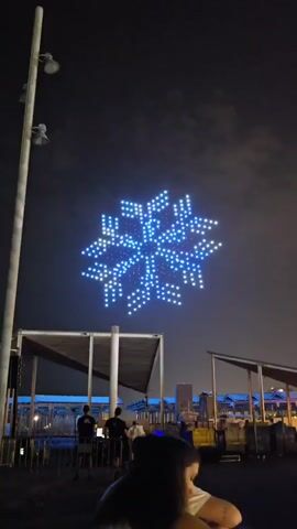 A drone light show features a snowflake-shaped formation of blue and white lights against a dark sky. In the foreground, silhouetted figures stand near a metal fence and a covered structure with visible beams. The overall scene appears to be an outdoor nighttime event.