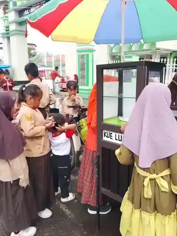 The video shows a wet, paved outdoor area in front of a building marked "AL-MUTTAQIEN". Several children in school uniforms are gathered around food vendors. One vendor is selling "KUE LE". There are several parked motorcycles and scattered pieces of paper on the ground. The scene suggests a school dismissal time with children buying snacks from street vendors. The weather is overcast.