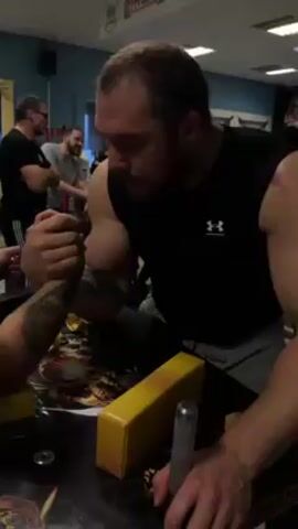 Here is a detailed description of the video:

The video shows a close-up of two men arm wrestling. The man closest to the camera is wearing a black Under Armour tank top and grey pants. He has short brown hair, a beard, and muscular arms. His opponent, seen partially, has tattoos on his left arm. They are arm wrestling on a table with yellow padding.  A silver metal pin sits to the right of the frame. The table has a black and gold patterned covering.

In the background, several other men are standing around watching and talking, some are smiling. They are wearing casual clothing. The room is brightly lit with fluorescent ceiling lights. There are posters on the wall, and overall the space appears to be a casual recreational area or gym. There are several tables with yellow pads on them, suggesting this is a dedicated space for arm wrestling. The camera slightly moves to capture more of the surrounding activity.