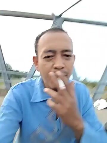 A man is sitting on a motor scooter on a bridge, facing the camera. He is wearing a light blue shirt with dark blue accents. The bridge has a metal frame structure above it. The man is looking at the camera. The background shows a landscape with vegetation and a road.