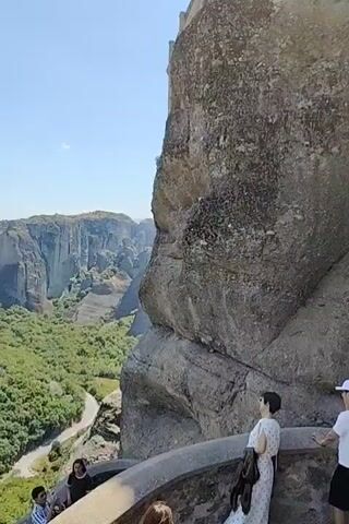 The video shows a panoramic view of Meteora in Greece. The camera pans across vast rock formations covered in lush green vegetation. In the distance, several monasteries are visible perched atop the rock spires. The sky is clear and blue with scattered white clouds. Several people are seen in the foreground, taking in the breathtaking scenery. One woman in a white dress is looking out at the view. A couple is standing on a balcony overlooking the valley. Another person is seen from behind, also enjoying the view. The video captures the majestic beauty of this unique natural and historical site.