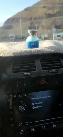 The video shows the interior of a car dashboard. A blue bottle of air freshener is on the dashboard. The car is moving, as indicated by the changing speed on the dashboard display, which shows 59, 62, and 66. The landscape outside the car window suggests a mountainous or hilly region with a road. In the background, there are vehicles, including trucks and vans, and power lines. The sky is blue and sunny. The video appears to be taken during daylight hours. The audio includes music.