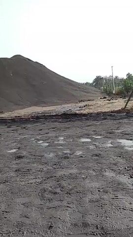 The video presents a static view of a landscape under an overcast sky. In the foreground, a muddy area is visible, marked with tire tracks and scattered puddles of water. In the background, a large, dark pile or mound rises against the horizon, resembling a hill or a mountain of material. To the right of the mound, a line of trees is visible, suggesting the presence of some vegetation. The video does not contain any discernable people or actions.