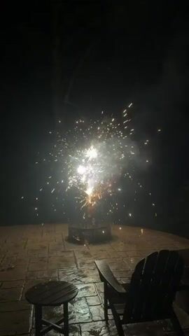 This is a video of a small firework exploding in a backyard at night. The firework is in a fire pit on a paved patio. There is a table and chairs in the foreground. The firework shoots sparks and smoke into the air.