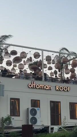 A low-angle, vertical shot captures an outdoor dining area on a rooftop during twilight. The building below has a white facade with three windows featuring wooden shutters and two air conditioning units mounted on the wall. Above the windows, a large sign reads "ottoman ROOF" in illuminated gold lettering. The rooftop itself is bustling with people seated at tables. Overhead, a decorative structure is adorned with numerous hanging lampshades made of woven natural materials, creating a warm and inviting ambiance. Palm trees are visible on the right side of the frame, silhouetted against the pale sky. The overall scene suggests a lively restaurant or bar setting in the evening.