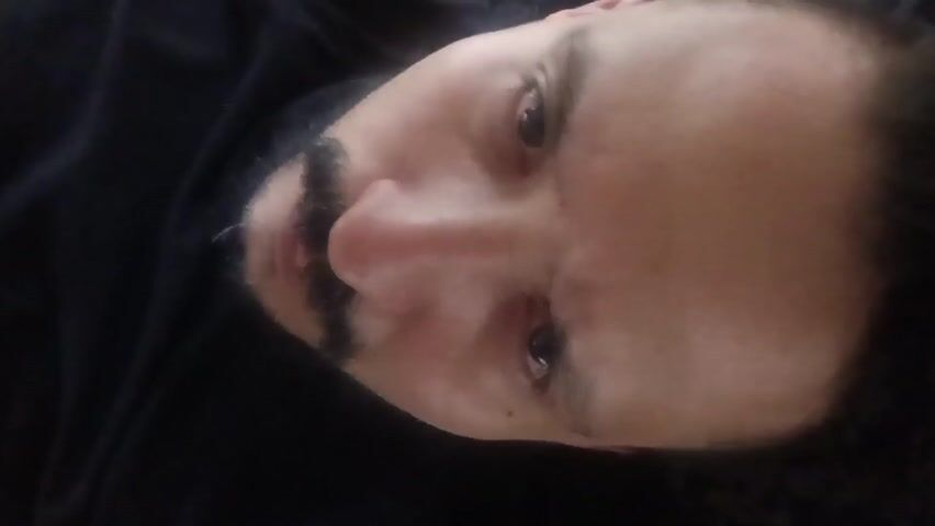A medium shot of a man's face, angled slightly downward. He has light skin, a trimmed dark mustache, and dark eyes. He is wearing a black hoodie, and the background is dark. He appears to be lying down. There are no visible actions or interactions in the frame.