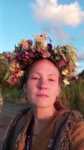 A first-person perspective video begins with a close-up of a woman wearing a large, colorful flower crown. Her expression is neutral as she looks directly into the camera. She is wearing a brown knitted sweater over a darker garment and a fur-lined jacket. A camera strap with the word "Canon" is visible around her neck. The background is slightly blurred, showing a dirt path and a field with trees. As the video progresses, the camera pans slightly to the right, revealing more of the environment and a gathering of people in the distance. The people appear to be outdoors, possibly at an event, with some individuals wearing flower accessories. The lighting suggests it is late afternoon or early evening, with the sun casting warm light. The scene seems festive and relaxed.