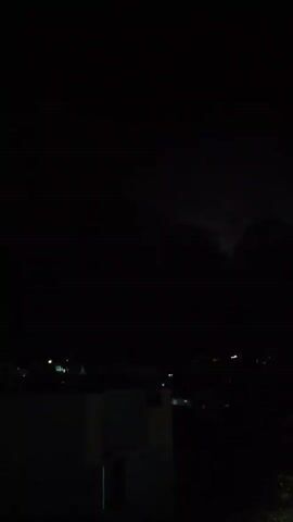 The video shows a night scene with a dark sky and a few faint lights scattered across a cityscape. The cityscape is viewed from an elevated position, with the rooftops of buildings visible in the foreground. The video is dark, and few details are discernible due to the lack of light. A bright flash of lightning is seen near the top of the frame at one point in the video.