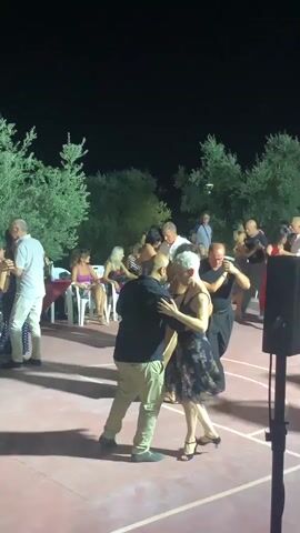 A group of people are dancing outdoors at night. There are trees and lights in the background. Several couples are dancing in the foreground, and others are watching from chairs or standing around. The ground is a reddish-brown surface, possibly a tennis court or basketball court. A black speaker is visible on the right side of the frame. The lighting suggests it is after sunset. The people are dressed in casual and semi-formal attire.