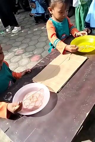 The video shows two young children, appearing to be girls, engaged in an activity outdoors. They are seated at a table. The girl on the right is wearing an orange and teal shirt and is holding a stick with what appears to be elastic bands on it. In front of her is a yellow bowl with more elastic bands. The girl on the left is wearing a green shirt and is also holding a stick in a pink bowl filled with elastic bands. A piece of cardboard is placed between them on the table. In the background, other people and children are visible, as well as motorcycles parked on a paved surface. The environment appears to be a sunny outdoor setting. The children seem focused on their task.